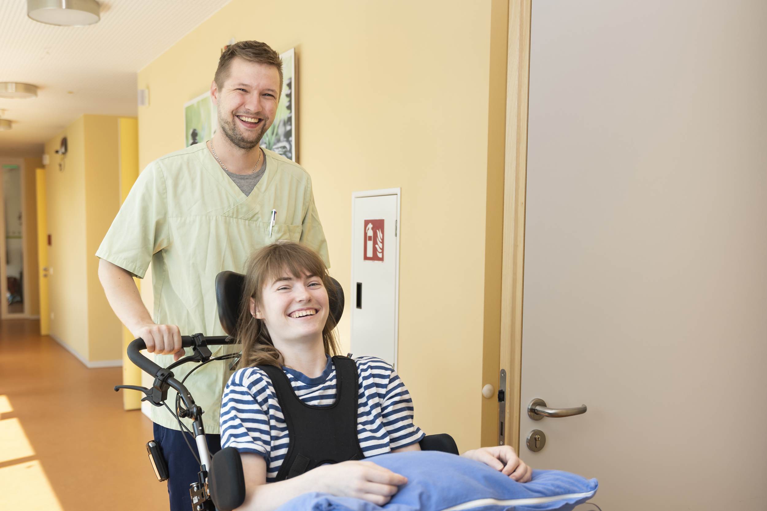Eine lächelnde VIVICARE Pflegekraft schiebt eine ebenfalls lächelnde Patientin in einem Rollstuhl durch einen Flur.