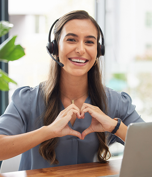 lächelnde Mitarbeiterin mit braunen Haaren, trägt ein Headset zum telefonieren und formt mit ihren Händen ein Herz