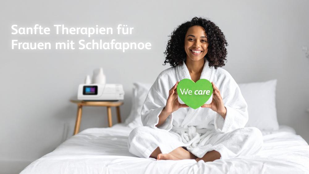 Eine junge, lächelnde Frau sitzt entspannt im weißen Bademantel auf einem Bett. Sie hält ein grünes Herz mit dem Text "We care" in den Händen. Im Hintergrund ist ein CPAP-Therapiegerät zu sehen. Das Motiv symbolisiert die sanfte, fürsorgliche Therapie für Frauen mit Schlafapnoe durch die VIVISOL Heimbehandlungsgeräte GmbH.