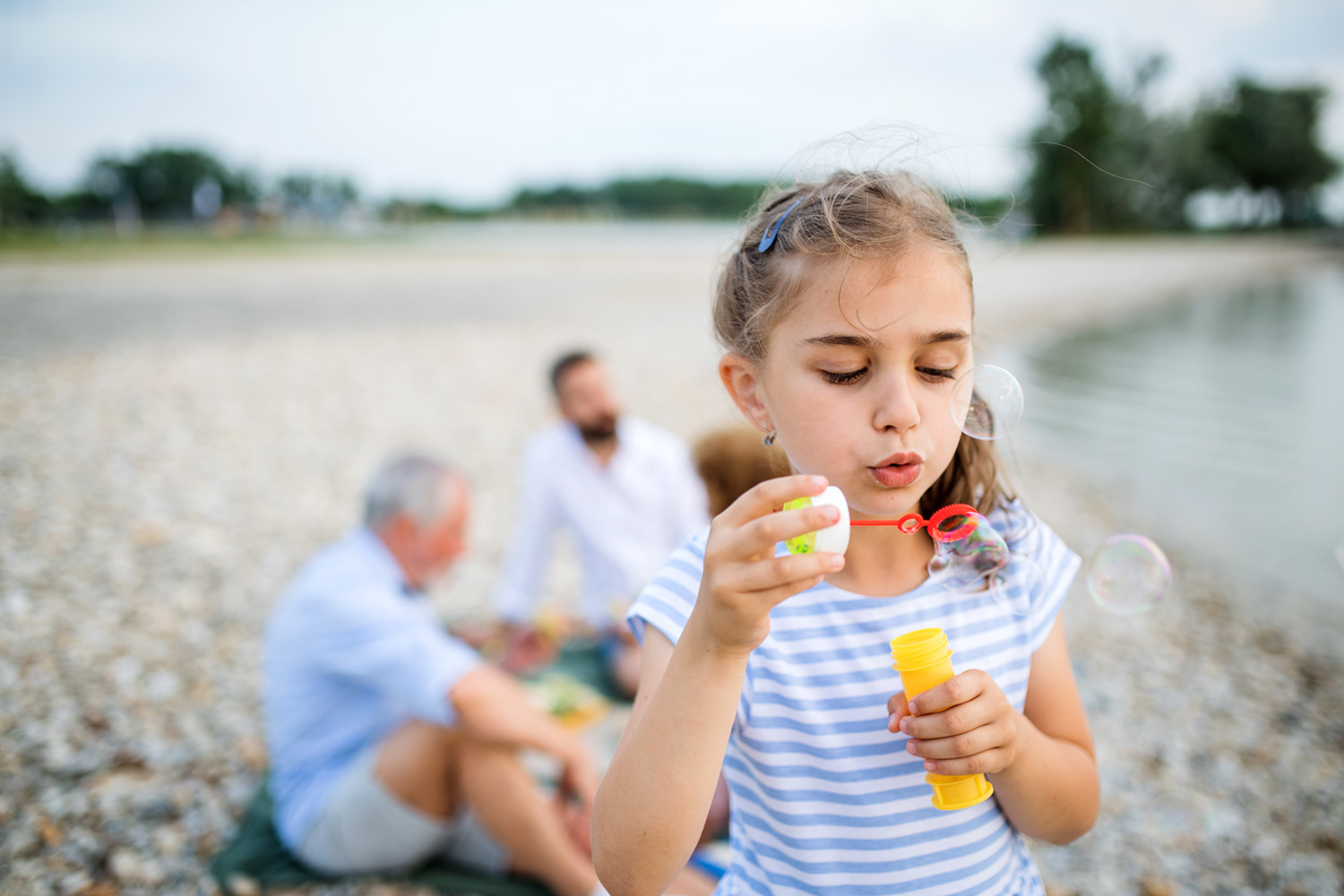 Ein Kind auf einem Kiesstrand, das mit geschlossenen Augen Seifenblasen bläst. Im Hintergrund sitzen erwachsene Familienmitglieder entspannt im Sand. Das Bild symbolisiert die erfolgreiche Sekretmobilisation, die Patienten (auch Kinder) freie Atemwege und die unbeschwerte Teilnahme am Familienleben ermöglicht.